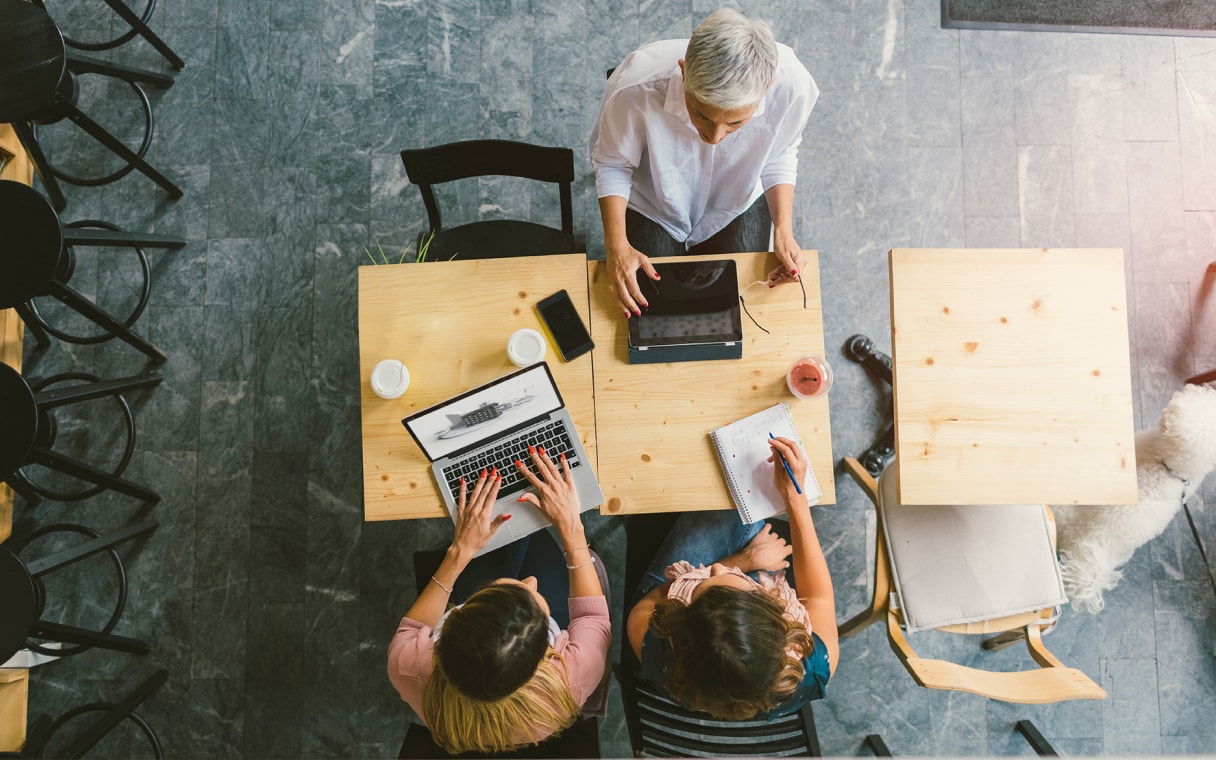 Three people sit at a table working on laptops and taking notes in notebooks.